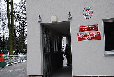 Zdjęcie kolorowe. Budynek usytuowany przy wejściu do Oddziału Prewencji Policji w Katowicach. Na budynku widoczne tablice urzędowe z nazwami instytucji i godło z orłem.