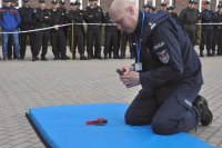 Policjant klęczy na materacu i składa rozłożony na części pistolet. W tle stoją inni policjanci.