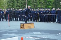 Policjant w biegu przeskakuje przez skrzynię gimnastyczną. W tle stoją inni policjanci.