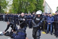 Trzech policjantów w trakcie zakładania sprzętu ochrony osobistej. Widać, że się spieszą. W tle stoją inni policjanci.