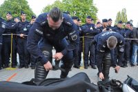 Dwóch policjantów w trakcie zakładania sprzętu ochrony osobistej. Widać, że się spieszą. W tle stoją inni policjanci.