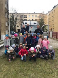 Grupa dzieci w wieku przedszkolnym oraz dwóch policjantów w mundurach pozują do zdjęcia na tle radiowozu.