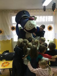 Zdjęcie w sali przedszkolnej. Maskotka policyjna Szupek otoczona i ściskana przez dzieci.