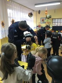 Policjant zakłada dziecku kask policyjny. Przyglądają się inne dzieci.