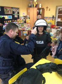 Chłopiec ubrany w kamizelkę kamienioodporną i kask policyjny, trzyma w rękach pałkę policyjną. Jest w towarzystwie innych dzieci i policjanta.