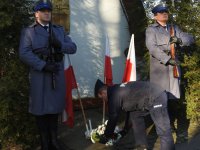 Zdjęcie kolorowe. Przed halą sportową policjant składa wieniec pod tablicą pamiątkową. Jest w asyście dwóch innych policjantów.