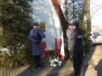 Zdjęcie kolorowe. Przed halą sportową policjant oddaje honor przed tablicą pamiątkową w asyście policyjnej dwóch policjantów.