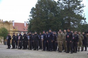 Policjanci w różnych mundurach stoją na zbiórce w kolumnach. Przed policjantami znaki identyfikujący przynależność.