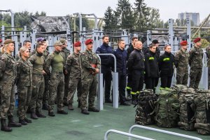 Osoby w mundurach wojskowych, policyjnych i straży pożarnej stoją w luźnym szyku. W tle maszyny do ćwiczeń.