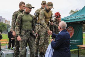 Osoby w zielonych mundurach stoją na podium. Mężczyzna w ubraniu cywilnym składa gratulacje.