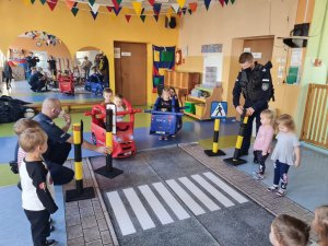 Policjanci podczas gry edukacyjnej, wyposażeni w rekwizyty takie jak mata imitująca ulicę, znaki drogowe i samochody, tłumaczą dzieciom zasady bezpiecznego zachowania się na drodze.