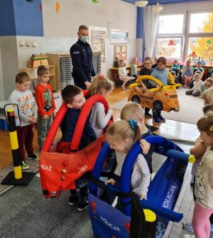 Policjanci podczas gry edukacyjnej, wyposażeni w rekwizyty takie jak mata imitująca ulicę, znaki drogowe i samochody, tłumaczą dzieciom zasady bezpiecznego zachowania się na drodze.