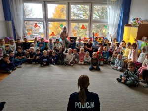 Grupa zaciekawionych przedszkolaków z uwagą słucha prelekcji wygłaszanej przez policjantkę.