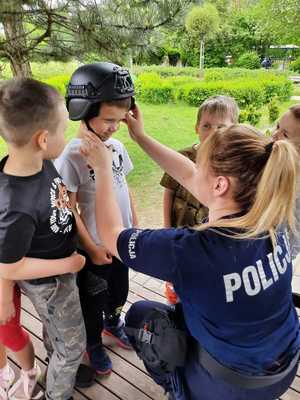 Policjanta pomaga chłopcu założyć hełm kevlarowy. Inne dzieci przyglądają się.