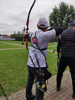 Zdjęcie. Sierżant Adam Ścibski podczas swojej próby trzyma łuk w lewej ręce, a prawą napina cięciwę z założona strzałą. Ujęcie od tyłu.