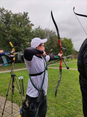 Zdjęcie. Sierżant Adam Ścibski podczas swojej próby trzyma łuk w lewej ręce, a prawą napina cięciwę z założona strzałą. Ujęcie z prawej tylnej strony.