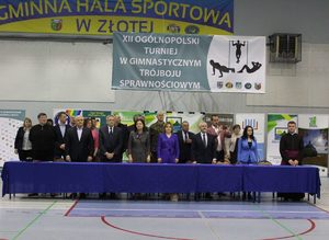 Organizatorzy zawodów i zaproszeni goście pozują do zdjęcia za stołem. Nad nimi wisi plakat z nazwą zawodów.