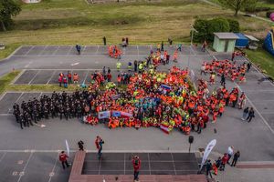 Zdjęcie z lotu ptaka. W dole grupa około 150 osób zgromadzona na placu. Ubrani w pomarańczowe stroje służb ratowniczych, mundury, żółte kamizelki. Kilka grup trzyma flagi.