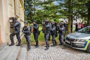 Grupa policjantów w rzędzie. Stoję w szyku przed drzwiami wejściowymi do budynku. Dwóch policjantów trzyma tarcze balistyczne.