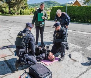 Na otwartym placu leży osoba w mundurze z widocznymi obrażeniami. Przy niej dwóch funkcjonariuszy. Widać torby medyczne. Obok dwóch obserwatorów.