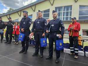 Na zdjęciu trzech policjantów w polskich mundurach. Mają na szyi medale. Zdjęcie na tle budynku. Obok klaszczące osoby.