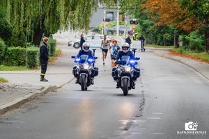Pierwsi trzej zawodnicy podczas biegu. Na pierwszym planie dwóch policjantów na motocyklach. Obok na chodniku strażak OSP.