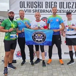 Pięciu policjantów Oddziału Prewencji Policji w Katowicach po zakończonym biegu pozuje z medalami i flagą z logo jednostki.