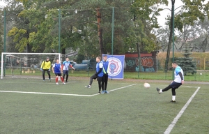 Zawodnik Kompanii Wzmocnienia oddaje strzał z rzutu wolnego przy boczne linii boiska. 2 zawodników drużyny przeciwnej w murze.