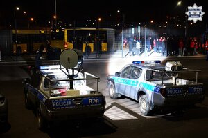 Dwa radiowozy z reflektorami oświetlają grupę kibiców pod stadionem.